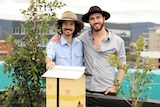 Connor and Jacob wear wide brimmed hats and smile in front of a native bee hive and two plants in a car park.
