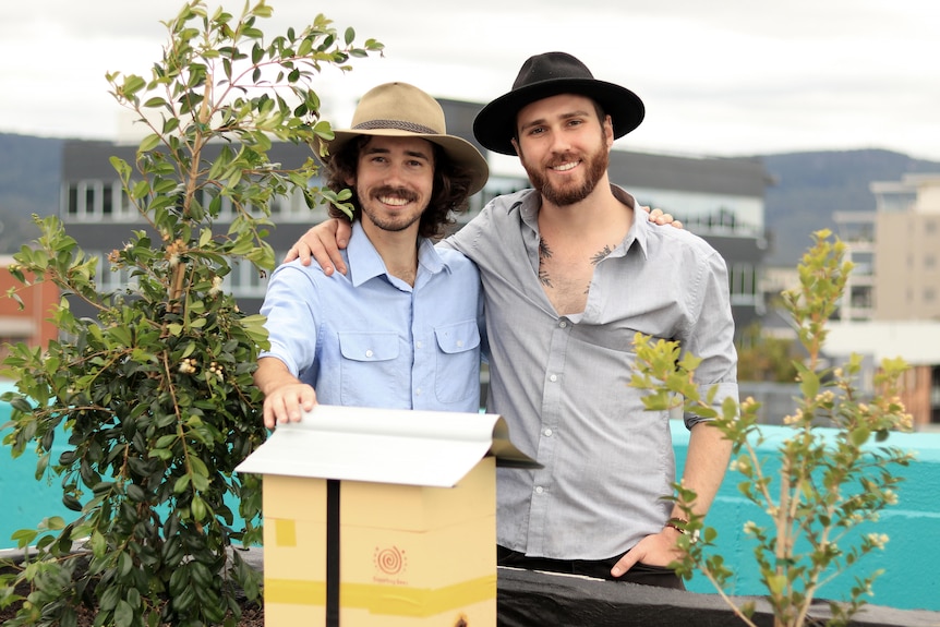 Connor and Jacob wear wide brimmed hats and smile in front of a native bee hive and two plants in a car park.