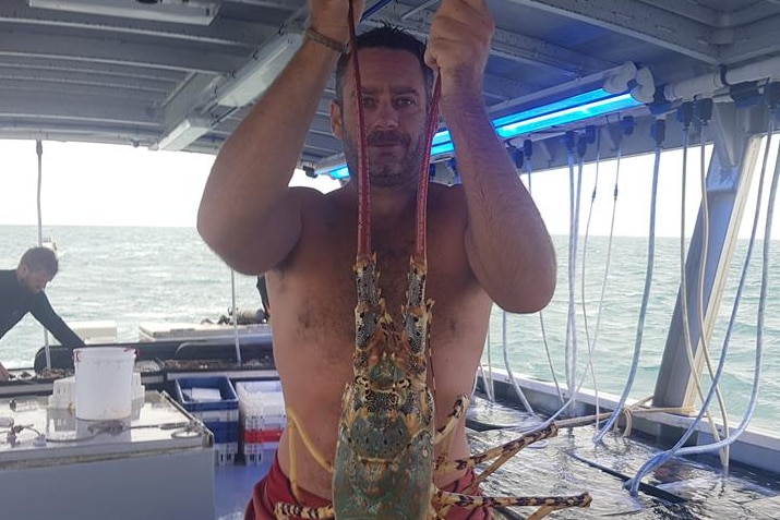 Joel Feeney standing on a boat deck holding up a huge tropical rock lobster.