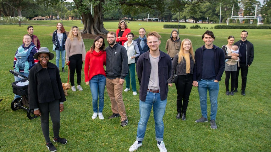 A group of people standing in a park with a man in jeans and a t-shirt out front.