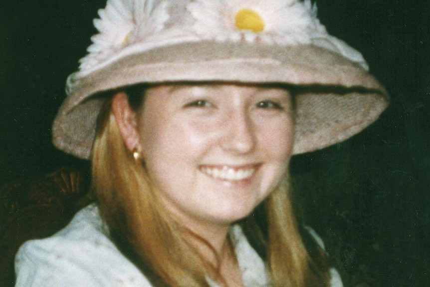 Sarah Spiers smiles for a photo sitting at a dinner table eating, wearing a straw hat with daisies on it and a blue shirt.