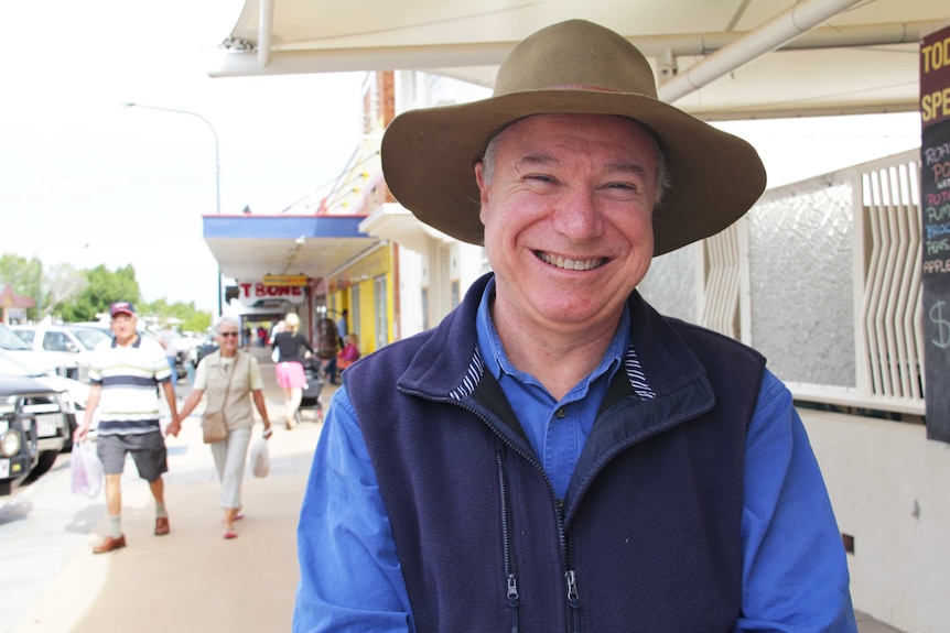 A man in a hat smiling at the camera with shoppers milling around the main street of a country town behind him.