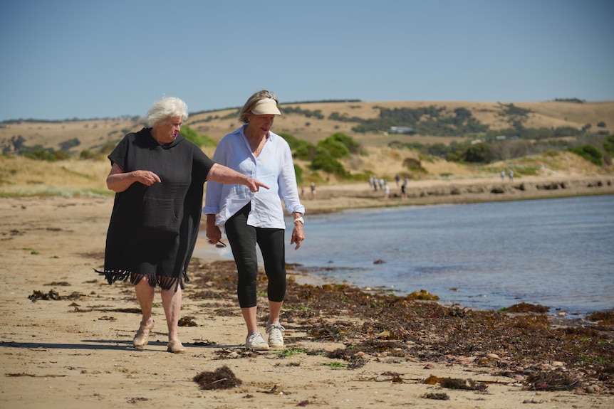 Two women walk along a beach at the edge of the water and near seaweed, one woman is pointing to something on the ground