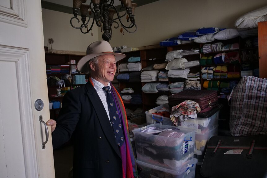 A man wearing an Akubra and rainbow scarf opens a door with helves full of linen and clothes behind him 
