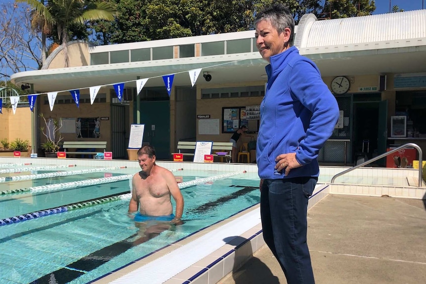 A man wades in waist-deep water in pool as woman watches on a bright and sunny day.