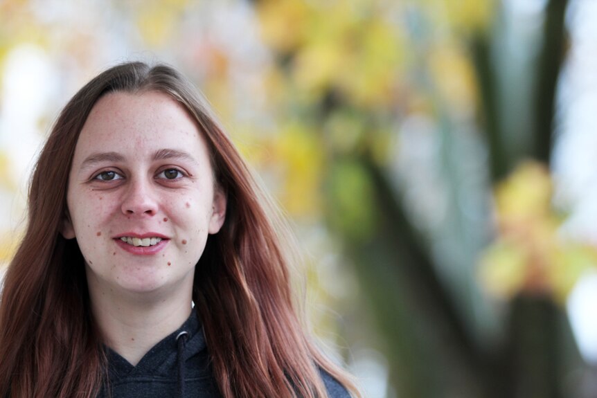 A young woman looks directly at the camera, with out-of-focus trees in the background.