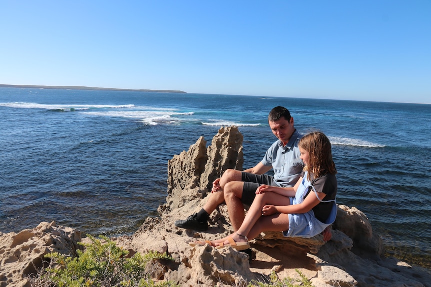 A man and a young girl wearing overalls sit on a rocky outcrop overlooking the ocean. The sky is clear and blue. 