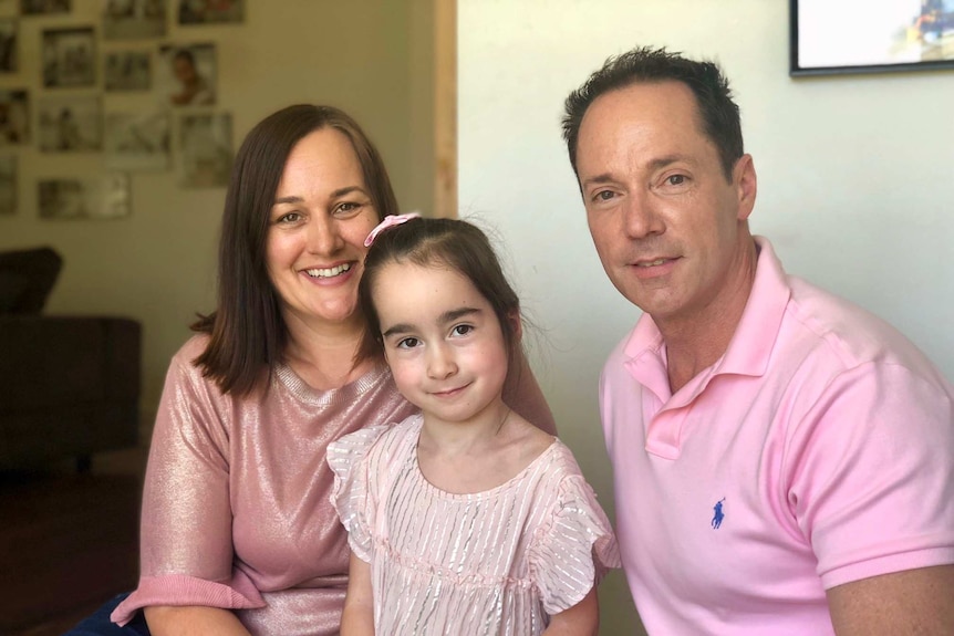 Two parents sit either side of their young daughter. The three of them are wearing pink, and the mother is smiling.