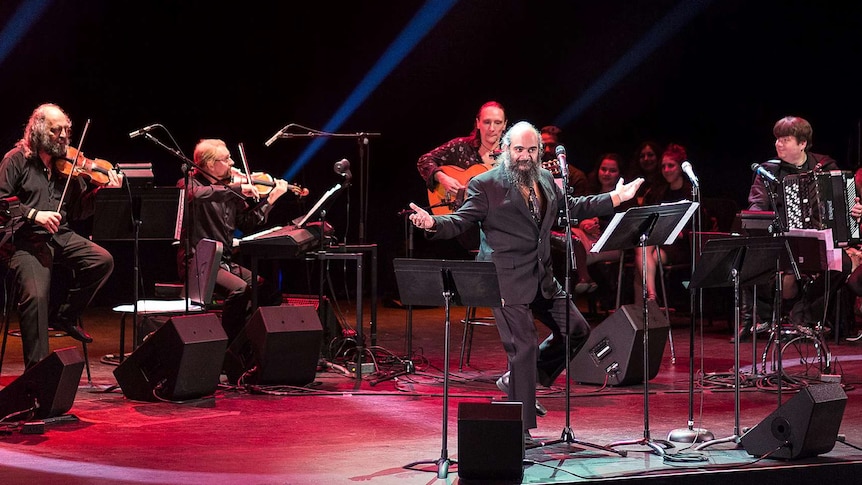 Two violinists, a guitarist and an accordion player accompany a singer standing at a microphone with his arms out.