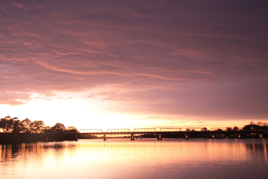 The sun is beginning to rise and the sky is orange. The Shoalhaven Bridge is silhouetted.