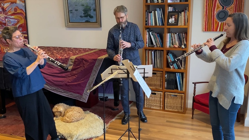 Three oboe players stand around a piano with a dog sitting under it