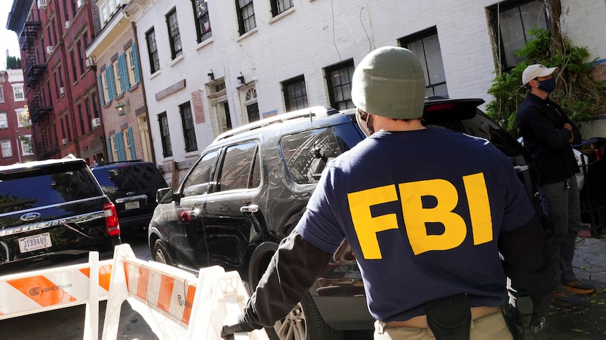 A photo of the back of a man standing on the street who has FBI on his vest in big yellow letter.