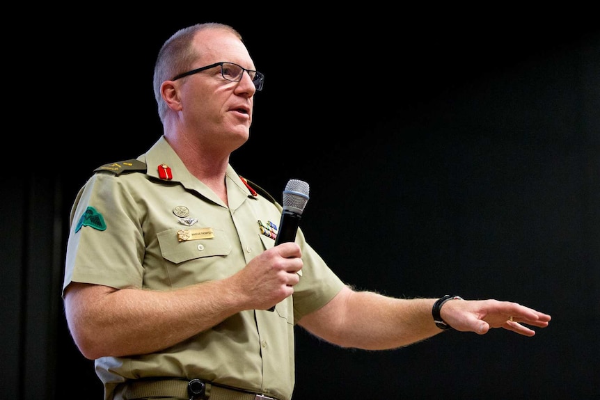 A man in military dress holds a microphone as he speaks at a meeting