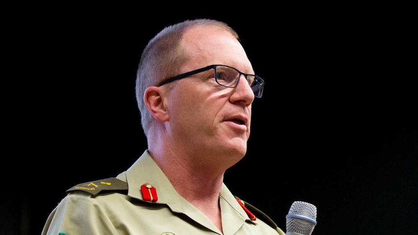 A man in military dress holds a microphone as he speaks at a meeting