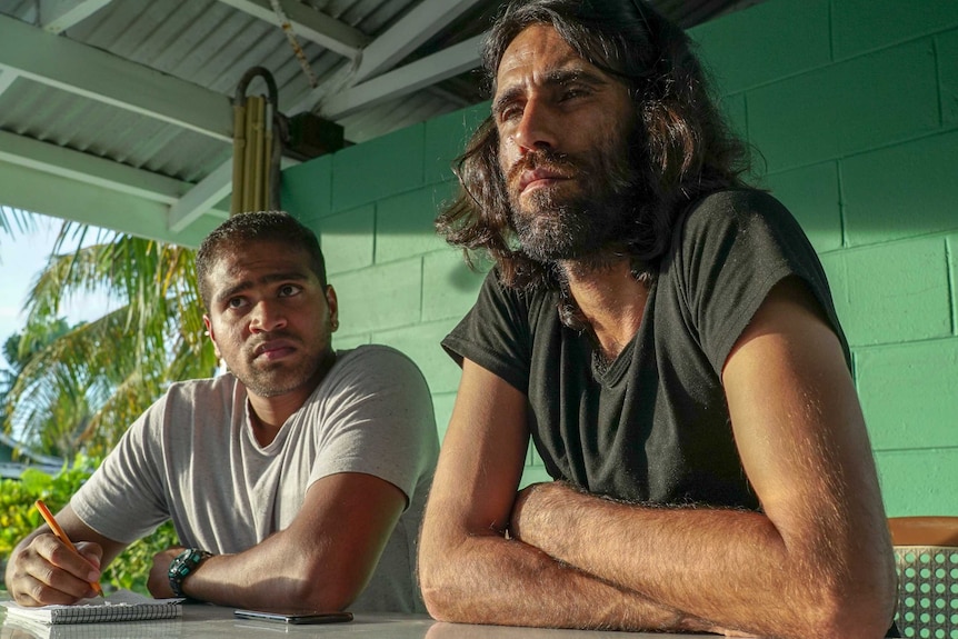 Two men looking serious while sitting at a table in a tropical location