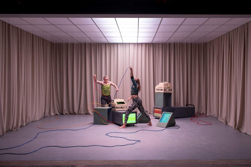 Small curtained stage with illuminated grid ceiling, grey office carpet, in which two dancers 'struggle' with TV-style objects.