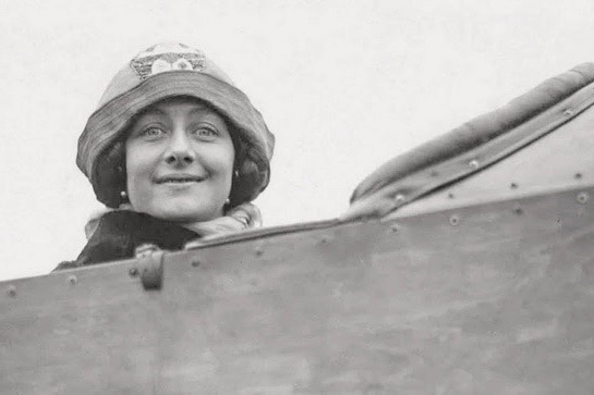 A woman with dark hair and wearing a hat sits in an aeroplane.
