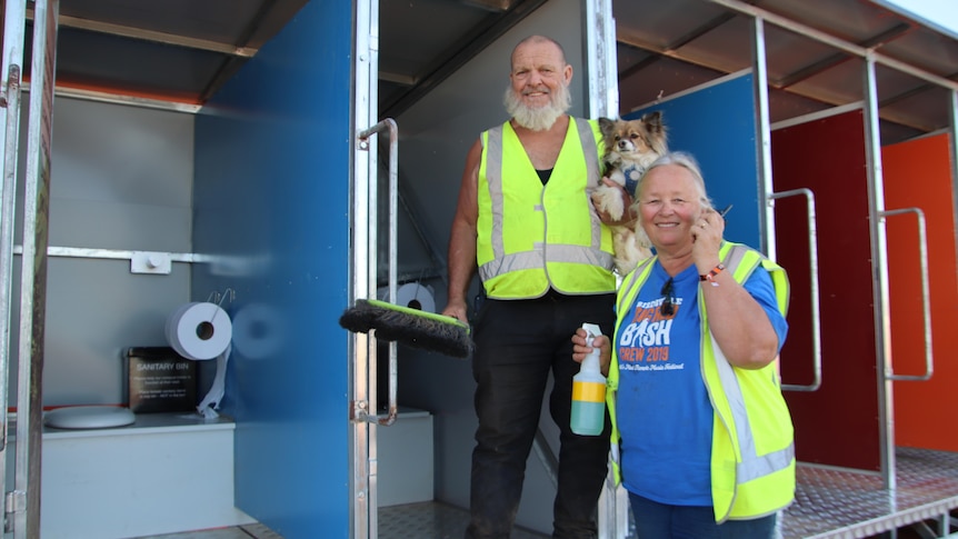 a man holding a dog and a broom and a woman holding a walkie talkie and spray bottle, both smiling, standing by toilet cubicles