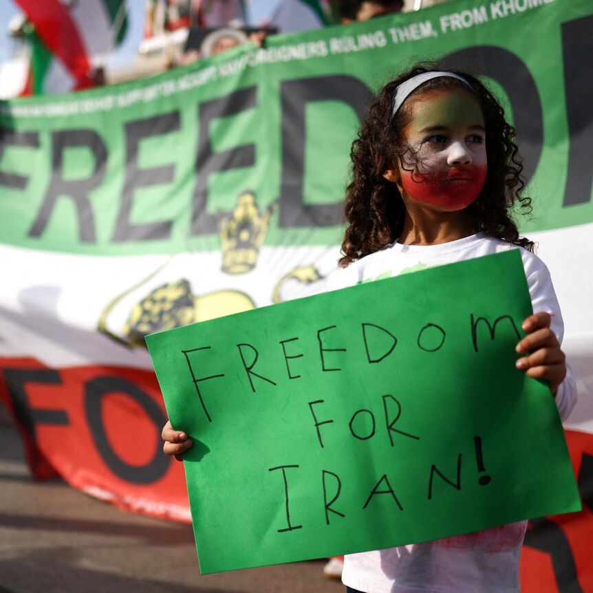 A little girl with her face painted in the colours of the Iranian flag holds a sign that reads FREEDOM FOR IRAN!