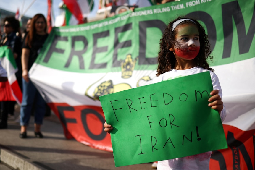 A little girl with her face painted in the colors of the Iranian flag holds a sign that reads FREEDOM FOR IRAN!