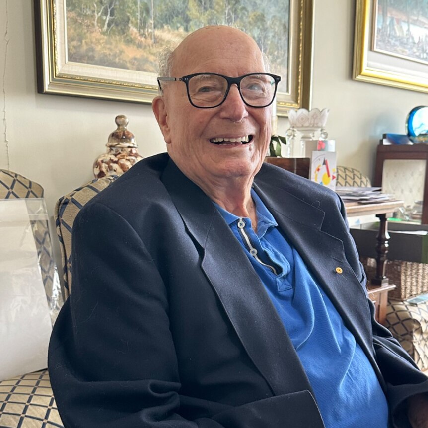 old man bald with spectacles smiling in a blue blazer in a loungeroom with gold framed paintings on wall