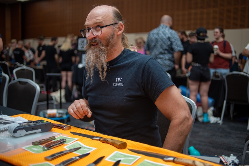 A man sits behind a table of knives.