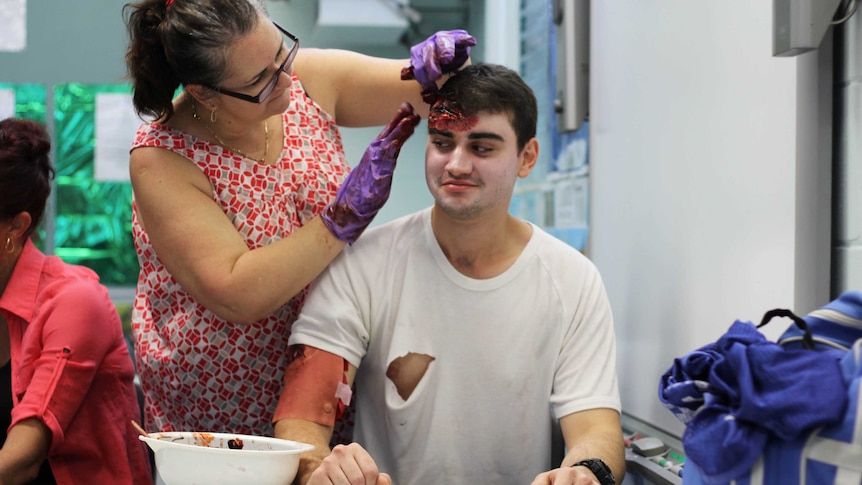 A young man sits at a table while a woman applies fake blood to his forehead.