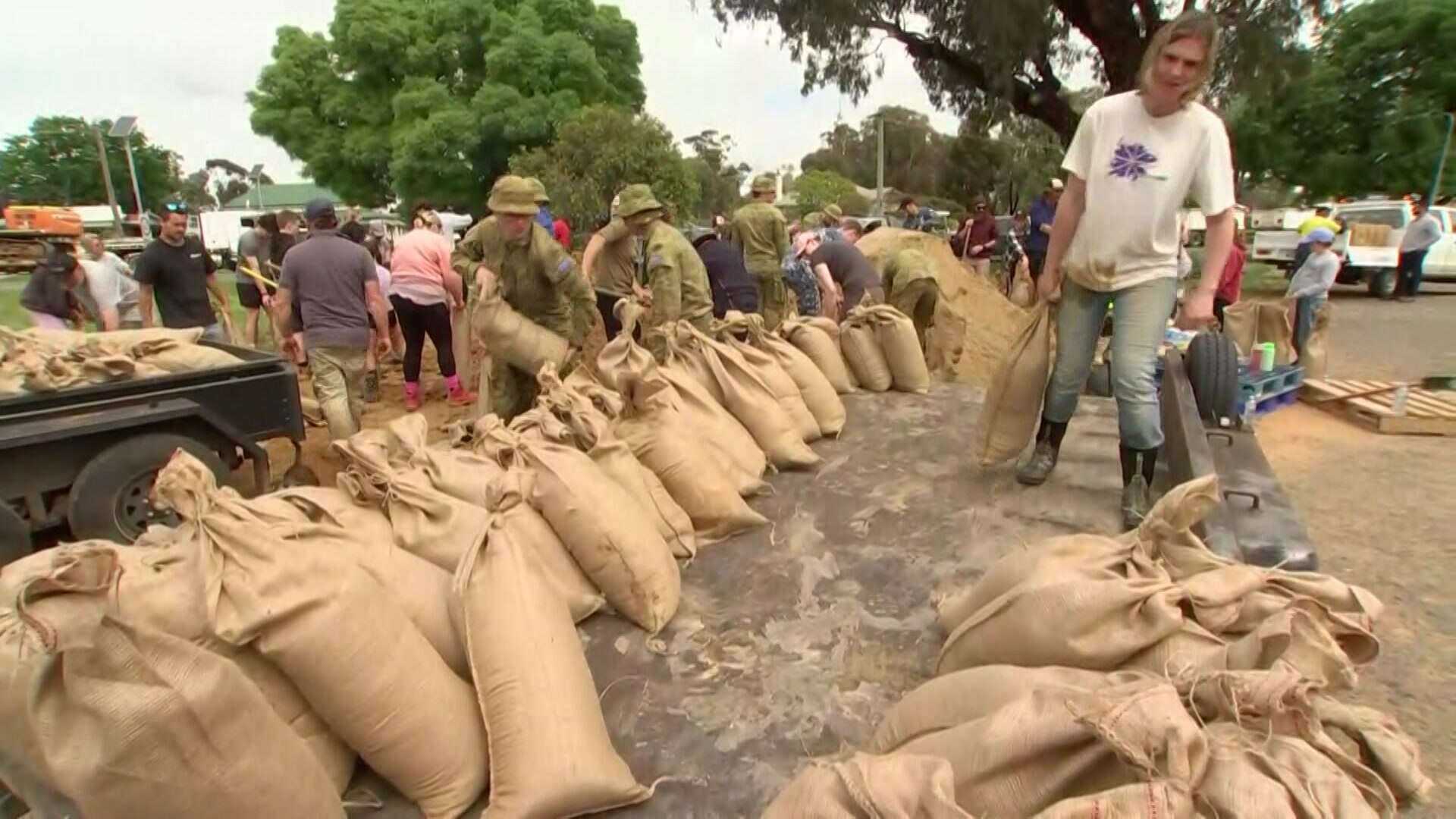 A line of full sandbags on a road with members of the defense force standing behind them and a man in carrying one.