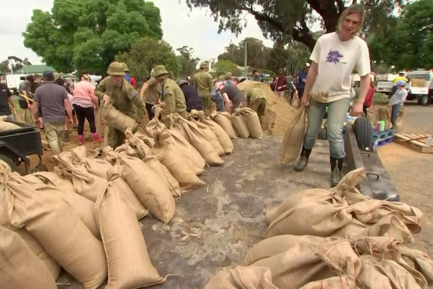 A line of full sandbags on a road with members of the defense force standing behind them and a man in carrying one.