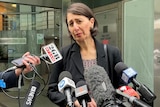 the former premier of new south wales gladys berejiklian stands outdoors behind microphones speaking to the media