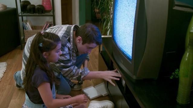 Teenage boy and young girl sit in front of TV, try to operate VCR