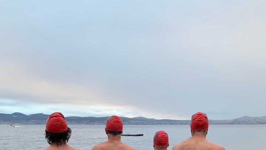 Four naked men wearing red swim caps stand around a fire pot with their backs to the camera.