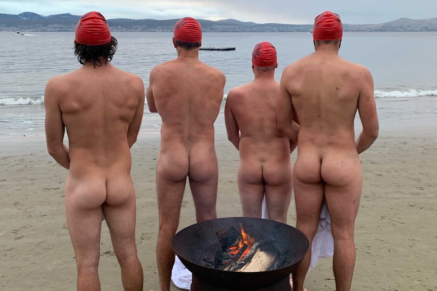 Four naked men wearing red swim caps stand around a fire pot with their backs to the camera.