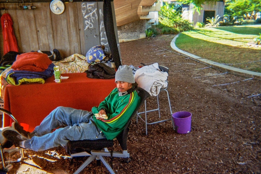 Homeless man sits eating a sandwich under a bridge.