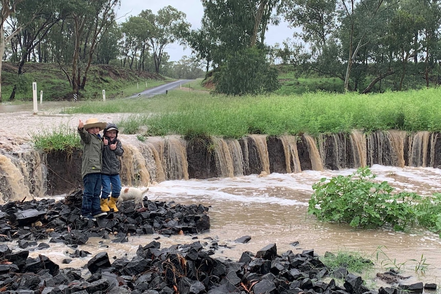 Two boys give the thumbs up standing beside a waterfall flowing into a creek after heavy rain