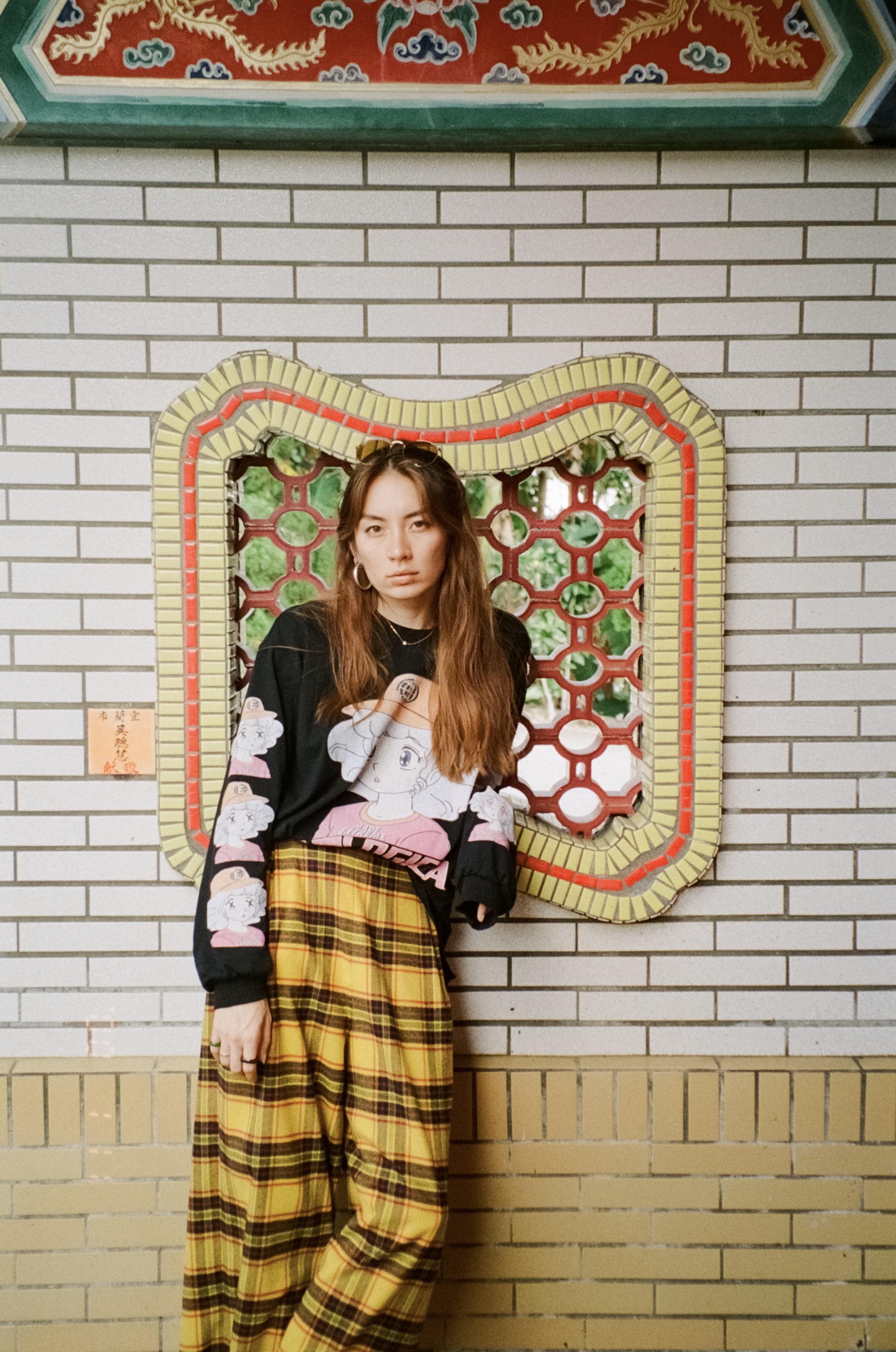 A Taiwanese Australian woman with long copper hair in an anime-print shirt and checkered pants leans against a wall in Taiwan.