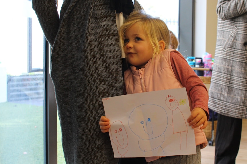A blonde haired child holds up a drawing of three stick figures