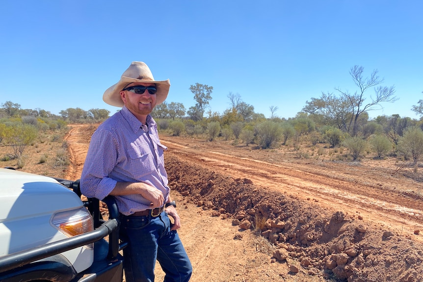 A man leans on the front of a white car in a paddock with banks of loose soil beside him