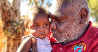 A man holds a baby who is wearing a singlet and nappy.