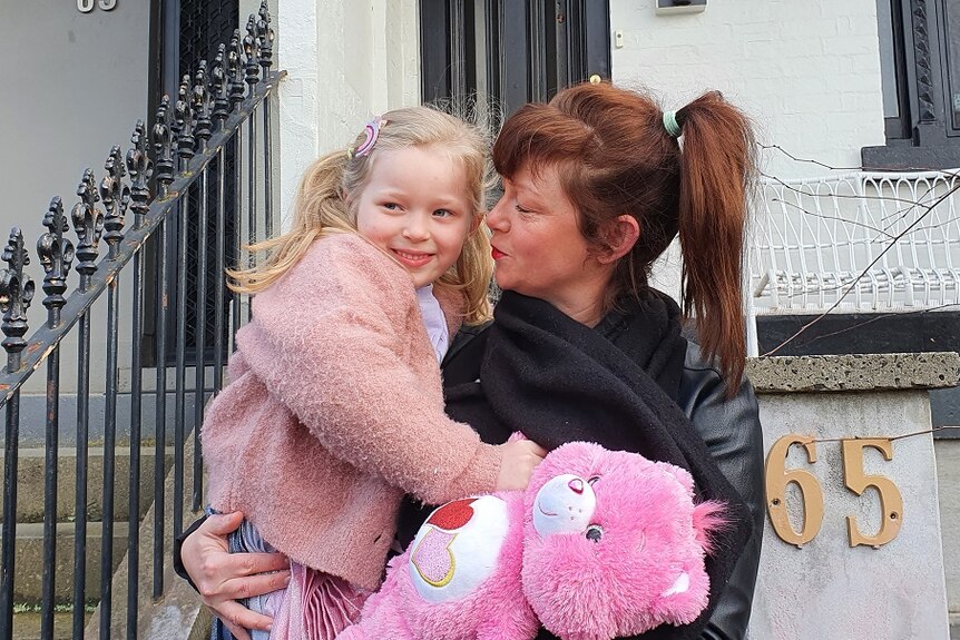 Caitlyn holds her smiling daughter Norah and a pink bear, in a urban residential street in Melbourne.