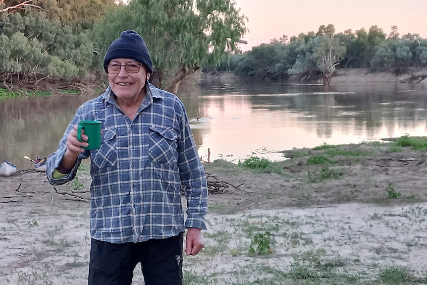 Man standing with long sleeve shirt, beanie and mug while smiling on river bank at sunrise