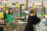 A young woman standing at a cash register looks at a woman sorting her groceries in a trolley.