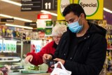 A man in a blue surgical mask opens his wallet at the checkout in Woolworths, while a woman behind him is not wearing a mask.
