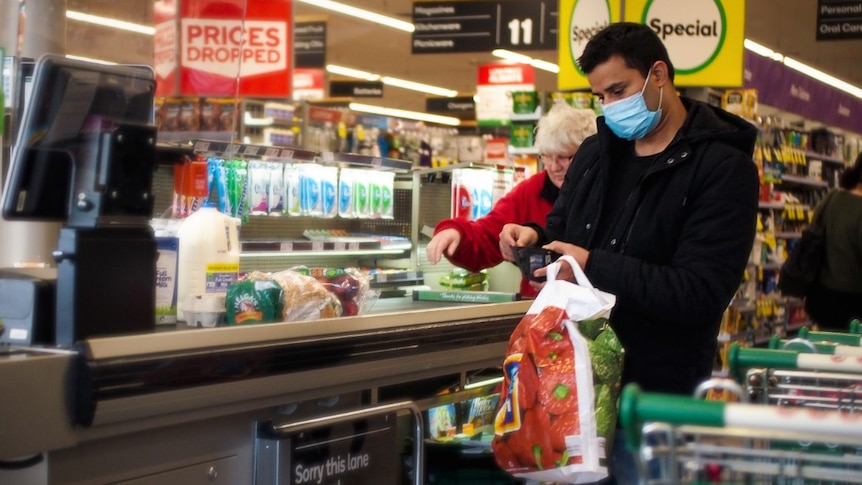 A man in a blue surgical mask opens his wallet at the checkout in Woolworths, while a woman behind him is not wearing a mask.