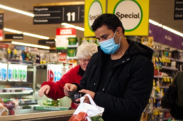 A man in a blue surgical mask opens his wallet at the checkout in Woolworths, while a woman behind him is not wearing a mask.