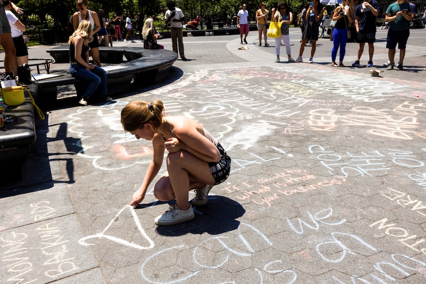 A woman writes on the ground, which is covered in writing, as observers watch on.