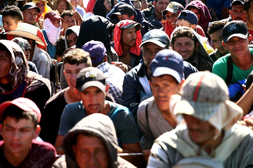 A group of migrants wearing hats and with t-shirts over the heads stand in a crowd with the sun on their heads
