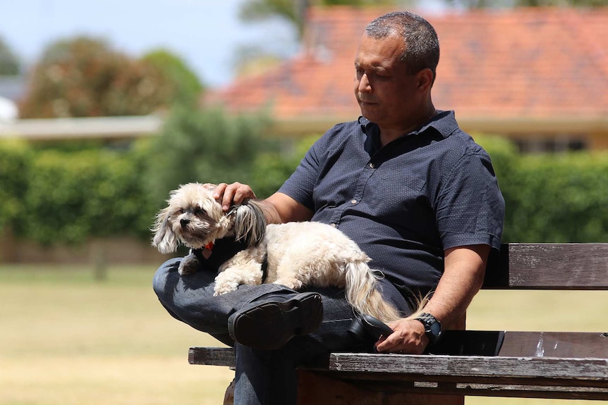 A man in a blue shirt sits on park bench with a small white dog on his lap.
