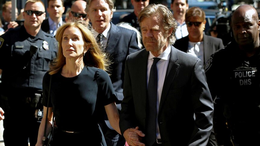A man and a woman, both wearing black business attire, hold hands as they walk into court with sombre expressions.