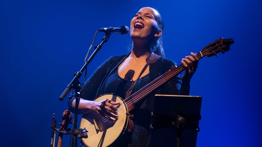 Rhiannon sings into a microphone in front of a blue background. She's holding a banjo.